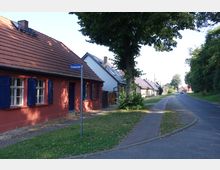Straßenansicht einer ländlichen Wohngegend mit kleinen Häusern, darunter ein rotes Gebäude mit blauen Fensterläden und einem Ziegeldach. Eine gepflasterte Gehwegfläche verläuft neben der Straße, die von Bäumen und Grünflächen gesäumt ist.