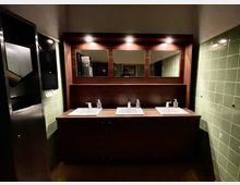 Waschraum mit drei weißen Waschbecken auf einer dunkelbraunen Holztheke. Darüber befinden sich drei beleuchtete Spiegel, und an den Seitenwänden sind grüne Fliesen angebracht.