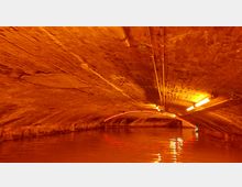 Ein unterirdischer Backsteintunnel mit gewölbter Decke, orangefarbener Beleuchtung und einem Wasserlauf, der den Boden des Tunnels bedeckt. Über der Decke verlaufen Rohrleitungen, und das Licht reflektiert auf der Wasseroberfläche.