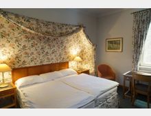 Ein gemütliches Hotelzimmer mit einem großen Doppelbett, flankiert von zwei Nachttischen mit Tischlampen. Die Wand hinter dem Bett ist mit einem floralen Stoff bespannt, der als dekoratives Element über das Bett drapiert ist. In der Ecke steht ein Sessel, daneben ein Schreibtisch mit Stuhl; ein Fenster mit floralen Vorhängen lässt Tageslicht herein.