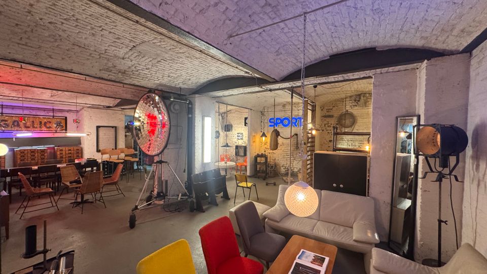 Industriewohnraum mit Gewölbedecke aus weiß gestrichenen Ziegeln, eingerichtet mit verschiedenen Sitzmöbeln, darunter bunte Stühle und Sofas. Auffällige Elemente sind ein großer Studioscheinwerfer, Neon-Schriftzüge an den Wänden und hängende Lampen; der Raum hat einen rustikalen und loftartigen Charakter.