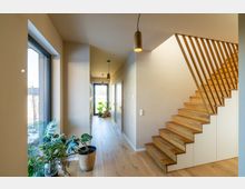 Moderner Wohnhausflur mit einer Holztreppe auf der rechten Seite, die mit vertikalen Holzstreben eingefasst ist, und einem hellen Korridor mit großen Fenstern und Zimmerpflanzen. Die Böden sind aus Holz, und die Beleuchtung erfolgt durch minimalistische Hängelampen.