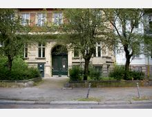 Fassade eines mehrstöckigen Altbaus mit großen, dunkelgrünen Doppeltüren und hohen Fenstern, umgeben von Bäumen und kleinen Grünanlagen, die teilweise durch niedrige Steinmauern eingefasst sind. Im Vordergrund befindet sich ein Gehweg und eine gepflasterte Zufahrt.