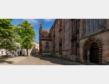 Backsteingotik-Kirche mit spitzbogigen Fenstern und filigranen Verzierungen, flankiert von einem gepflasterten Platz mit Bäumen und umliegenden Wohngebäuden. Im Vordergrund ist ein Eingang mit einem gemauerten Spitzbogen sichtbar.
