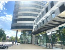 Ein modernes, mehrstöckiges Gebäude mit einer runden Fassade und Glasfront im unteren Bereich, daneben ein Nebengebäude mit rechteckigen Fenstern und Vordächern. Im Vordergrund eine breite, gepflasterte Gehfläche, auf der Personen und Fahrräder zu sehen sind, sowie einige Bäume und eine Straße im Hintergrund.