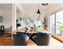 Essbereich in einem modernen, offenen Wohnraum mit einem Holztisch, gedeckt mit Geschirr und Gläsern. Im Hintergrund befindet sich eine grüne Wanddekoration mit Pflanzenmotiven, graue Möbel und große Fenster, die viel Tageslicht hereinlassen.