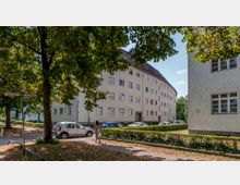 Wohnsiedlung mit schlichten Mehrfamilienhäusern in grauer Putzfassade, umgeben von Bäumen und Grünflächen. Im Vordergrund führt ein Gehweg entlang geparkter Autos, und Straßenschilder mit der Aufschrift „Am Fenn“ sind sichtbar.