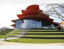 Moderne Veranstaltungsgebäude mit auffälligem geschwungenem, roten Dach und einer Fassade aus großen Glasflächen. Vor dem Gebäude befinden sich stufenförmig angelegte Grünflächen mit Treppen.