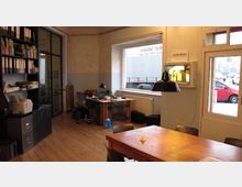 Ein einfacher Büroraum mit Holzboden, großen Fensterfronten und schlichten Möbeln. Im Vordergrund steht ein Holztisch mit Stühlen, im Hintergrund sind Schreibtische, Regale mit Ordnern und eine Glastür zu sehen.