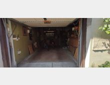 Offene Garage mit Fahrradstellplätzen und verschiedener Lagerung. Innen sind Regale, Werkzeuge und Holzmaterialien sichtbar, mit einer sichtbaren Schwingtür-Decke.