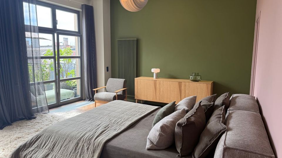 Modern bedroom with a large upholstered bed and layered pillows, a green accent wall with a tall radiator and wooden sideboard, and a round pendant light overhead. Floor-to-ceiling glass doors with sheer curtains open to a small balcony with plants, bringing in natural light.