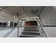 Innenansicht eines modernen Gebäudefoyers mit einer breiten Treppe aus grauem Stein und Edelstahlgeländern, die zu einer erhöhten Plattform führt. Darüber eine geneigte, paneelartige Decke mit mehreren Leuchten; links große Fenster und am Ende eine Holzverkleidung mit Türen.