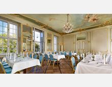 Barockes Speisezimmer mit reich verzierten Wänden und Decken, goldenen Ornamenten, einem Kronleuchter und bemaltem Deckengemälde. Der Raum ist mit weiß gedeckten Tischen, blau-goldenen Stühlen und großen Fenstern ausgestattet, durch die Tageslicht und ein Blick auf den Berliner Fernsehturm sichtbar sind.