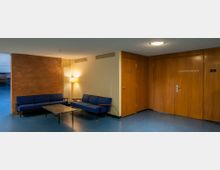 Sitzbereich mit zwei blauen Sofas und einem Glastisch in einer Ecke eines Gebäudes. Die Wände sind teilweise mit Holz verkleidet, mit Türen, die zu „Konferenzraum 3“ und einem Technikraum führen, sowie einer beleuchteten Lampe neben einem weißen Heizkörper.