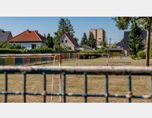 Ein Fußballplatz mit mehreren Torrahmen auf einer trockenen Wiese, umgeben von einem Maschendrahtzaun. Im Hintergrund sind Wohnhäuser mit roten und grauen Dächern sowie ein mehrstöckiges Gebäude und Bäume zu sehen.