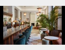 Innenansicht einer stilvollen Bar mit einer langen Theke aus Metall, daneben gepolsterte Barhocker in Blaugrün. Im Hintergrund befinden sich Sitzbereiche mit kleinen Tischen, Polstermöbeln und üppigen Zimmerpflanzen. Die Decke ist hoch mit dekorativen Hängelampen, der Boden ist mit einem gemusterten Fliesenmuster gestaltet.