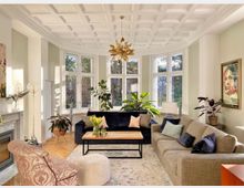 Wohnzimmer mit großen, gewölbten Fenstern, einer Kassettendecke und viel Tageslicht. Die Einrichtung umfasst eine graue und eine dunkelblaue Couch mit farbigen Kissen, einen Sessel mit gemustertem Stoff, einen Couchtisch aus Holz, Zimmerpflanzen und einen Kamin mit Marmorumrahmung.