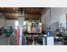 Werkstatt mit alten landwirtschaftlichen Maschinen und Geräten. In der Mitte steht ein großer, abgenutzter gelber Traktor; umgeben von Werkzeugen, Gasflaschen und Werkbänken vor Wänden aus weißem Mauerwerk und einem hölzernen Dachstuhl.