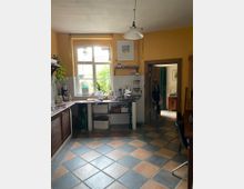 Eine gemütliche Küche mit gelb gestrichenen Wänden und einem großen Fenster, das Tageslicht hereinlässt. Der Raum ist mit einer Arbeitsplatte, einer Spüle, Regalen und Haushaltsgegenständen ausgestattet, der Boden ist mit farbigen Fliesen in einem Rautenmuster ausgelegt.