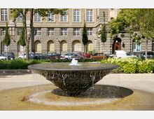 Ein runder Brunnen mit flachem, schalenförmigem Design, aus dem Wasser fließt, befindet sich in einer Pflasterfläche. Im Hintergrund steht ein großes historisches Gebäude mit symmetrischen Fenstern und Fassadenelementen aus Stein, umgeben von Bäumen, Blumenbeeten und geparkten Autos.