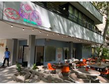 Außenbereich eines Cafés mit runden Tischen und orangefarbenen Stühlen, umgeben von Betonpflanzkübeln mit kleinen Pflanzen. Über dem Eingang ist ein bunter Neonschriftzug mit einer lächelnden Katze zu sehen, und Lichterketten sind an der Decke befestigt.