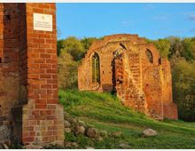 Ruine einer historischen Backsteinkirche in einer ländlichen Umgebung, umgeben von grünem Gras und Bäumen. Die Kirche hat teilweise erhaltene Wände mit gotischen Fensterbögen, und eine Informationstafel ist an einem der Backsteinelemente angebracht.