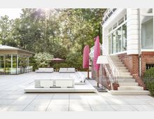 Heller Außenbereich mit moderner Terrasse, großen weißen Sitzmöbeln und rosa Sonnenschirmen, umgeben von Bäumen und Sträuchern; rechts eine Treppe, die zu einem Gebäude mit weißen Fensterrahmen und roter Ziegelfassade führt. Links ein verglaster Anbau mit Barhockern.