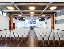 Großer Konferenzraum mit zahlreichen weißen Stühlen in Reihen auf einem dunklen Holzboden, einem Podium mit fünf schwarzen Stühlen und einer großen Leinwand im Hintergrund. Die Decke besteht aus Holzpaneelen mit runden Deckenlampen, und die Wände haben große Fenster und schlichte Dekoration.