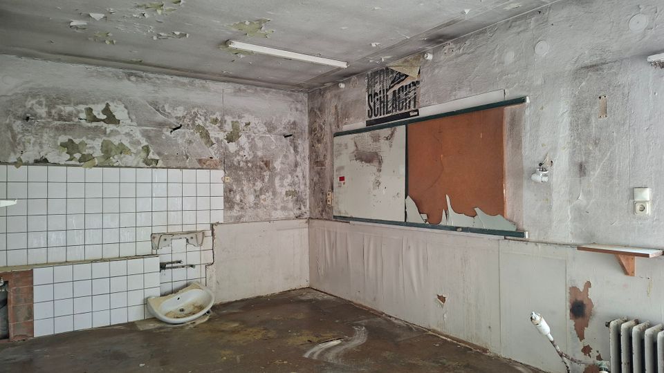 Verlassener Innenraum mit stark abgeplatzter Farbe und feuchten, fleckigen Wänden, teils mit weißen Wandfliesen und einem beschädigten Waschbecken. An der Decke hängt eine Leuchtstoffröhre; an der Wand sind ein leerer Schaukasten sowie ein alter Heizkörper und ein kleines Regal zu sehen.