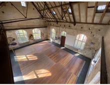 Großer, offener Raum mit hohen Decken und Holzboden, umgeben von unverputzten Backsteinwänden mit großen, bogenförmigen Fenstern. An den Wänden befinden sich farbige Graffiti, und das Dach zeigt freiliegende Holzbalken und Metallträger.