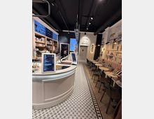 Ein modernes Eiscafé mit geschwungenem, weißem Tresen, auf dem Speisekarten stehen. Links befinden sich Regale mit Produkten, rechts Sitzbereiche mit gepolsterten Bänken, kleinen Holztischen und schwarz-gerahmten Stühlen. Die Wände zeigen kunstvolle Verzierungen und Motive, der Boden ist mit gemusterten Fliesen gestaltet.