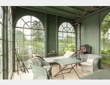 Grüner Wintergarten mit großen, bogenförmigen Fenstern, die den Blick auf eine Landschaft mit Bäumen und Schilf bieten. Der Raum ist mit geflochtenen Sesseln, einem Tisch und einer Liege ausgestattet; an der Wand befindet sich ein Kaminrohr.