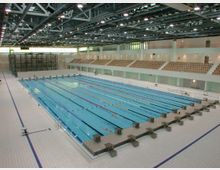 Innenansicht eines modernen Schwimmbads mit einem großen olympischen Schwimmbecken, das in mehrere Bahnen unterteilt ist, umgeben von einer weißen, gefliesten Fläche. Im Hintergrund befinden sich Zuschauertribünen mit Holzbänken sowie hohe Fenster und eine Dachkonstruktion mit sichtbaren Stahlträgern.