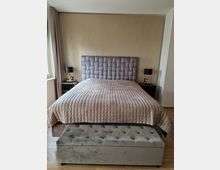 Schlafzimmer mit einem großen Bett mit gesteppter Kopfstütze und beige-gemustertem Überwurf, flankiert von zwei schwarzen Nachttischlampen auf dunklen Nachttischen. Vor dem Bett steht eine gepolsterte, graue Sitzbank, und der Raum hat Parkettboden.