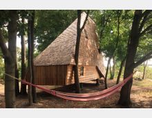 Ein ungewöhnliches Holzhaus mit steilem, asymmetrischem Dach steht auf einer Lichtung im Wald. Im Vordergrund ist eine rot-gestreifte Hängematte zwischen zwei Bäumen gespannt.