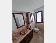 Badezimmer mit natürlichem Stil: Holzwaschbecken auf einer Steinplatte, darüber ein großer Holzrahmenspiegel; beige Wand- und Bodenfliesen, ein wandhängendes WC, ein Fenster mit dunklem Holzrahmen und Dekorationen wie Trockenblumen und Flaschen.