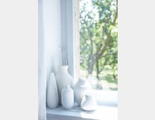 Eine Fensterbank mit mehreren weißen, strukturierten Vasen vor einem großen Fenster, durch das eine sommerliche grüne Landschaft mit Bäumen zu sehen ist.