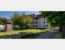 Wohngebäude aus den 1920er Jahren mit heller Fassade, rotem Satteldach und dekorativen Backsteinelementen, umgeben von einem grünen Rasen mit Blumenbeeten, einem Baum und einem Denkmal aus Stein. Rechts führt ein gepflasterter Gehweg entlang von Bäumen.