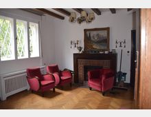 Wohnzimmer mit Holzboden und Balkendecke, ausgestattet mit roten Polstersesseln, einem Kamin mit gemauerter Verkleidung und darüber einem gerahmten Gemälde. Ein großes Fenster mit Gittermustern bietet Blick ins Grüne.