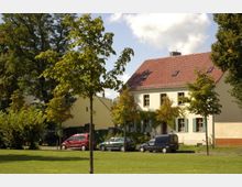 Ein zweigeschossiges Wohnhaus mit hellgelber Fassade und rotem Ziegeldach, umgeben von Bäumen und einer Grünfläche im Vordergrund; mehrere parkende Autos stehen entlang der Straße vor dem Gebäude.