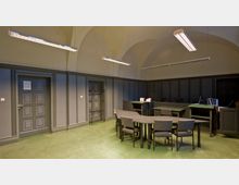 Ein schlichter Innenraum mit gewölbter Decke, grau gestrichenen Wandpaneelen und einem grünen Boden. Im Vordergrund steht ein dunkler Tisch mit mehreren Stühlen, während im Hintergrund Türen, ein Garderobenständer und ein Schreibtisch mit Büromaterialien zu sehen sind.