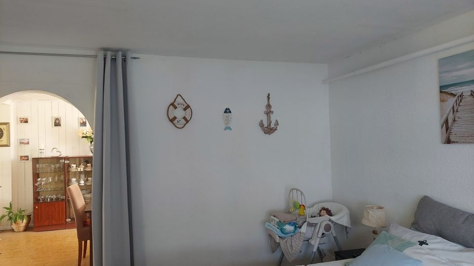 Heller Wohnraum mit weißen Wänden, maritimer Wanddekoration und einem Bett im Vordergrund; daneben stehen ein Babyhochstuhl und ein Nachttisch mit Lampe. Links führt ein Rundbogen in einen angrenzenden Essbereich mit Vitrine und Stühlen, getrennt durch einen grauen Vorhang.