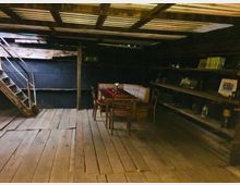 Holzinterieur eines rustikalen Raumes mit Holzboden, einer Metalltreppe auf der linken Seite, Wandregalen mit verschiedenen Gegenständen auf der rechten Seite und einem Tisch mit Stühlen in der Mitte. Über dem Raum befindet sich eine einfache Dachkonstruktion mit Wellblech.