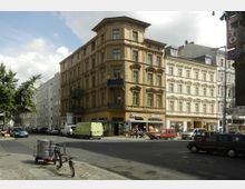 Ecke einer Stadtstraße mit einem vierstöckigen, historischen Gebäude in beige-brauner Fassade und reich verzierten Fenstern. Im Erdgeschoss befinden sich Geschäfte, während auf der Straße parkende Autos, ein Ladekarren und ein Fahrrad sichtbar sind; daneben stehen moderne, schlicht gestaltete Gebäude.