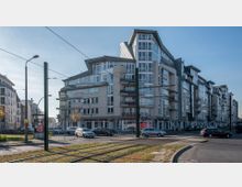 Straßenszene mit modernen, mehrstöckigen Wohn- und Geschäftshäusern, die helle Fassaden und große Fenster aufweisen. Im Vordergrund befinden sich Straßenbahnschienen, parkende Autos und Fußgängerbereiche, umgeben von vereinzelten Bäumen.