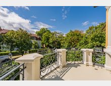 Blick von einem hellen Balkon mit dekorativem schmiedeeisernem Geländer und massiven Eckpfeilern auf eine von hohen Bäumen gesäumte Wohnstraße. Im Hintergrund sind mehrstöckige Wohnhäuser mit roten Ziegeldächern unter blauem Himmel zu sehen.