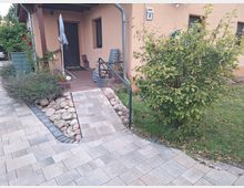 Privater Eingangsbereich eines Hauses mit beigem Putz, einem gepflasterten Zugang und einer kleinen Rampe. Neben der Rampe befinden sich ein Busch, Steine, Lavendel und weitere Pflanzen; im Hintergrund eine Terrasse mit Sitzmöbeln und einem Sonnenschirm.