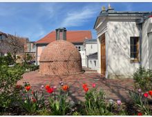 Garten mit blühenden roten und pinken Tulpen im Vordergrund, neben einer runden, kuppelartigen Backsteinstruktur. Im Hintergrund stehen Gebäude mit roten Ziegeldächern und weißer Fassade unter blauem Himmel.