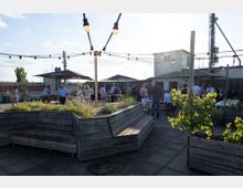 Dachterrasse mit Holzsitzbänken und Pflanzkästen, beleuchtet von dekorativen Lichterketten. Mehrere Personen stehen und sitzen an Tischen unter Sonnenschirmen, während Gebäude und eine Antennenanlage im Hintergrund zu sehen sind.