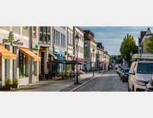 Straßenszene mit gepflastertem Weg, modernen Gebäuden mit bunten Fassaden und Geschäften sowie Restaurants mit Sonnenschirmen und Markisen; geparkte Autos und Laternen säumen die Straße, im Hintergrund sind Bäume sichtbar.